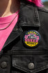 Round pinback button badge with Local Queer Menace text in bold yellow and pink retro lettering on a black background. A cartoon raccoon with a spiked collar flips the middle finger with a painted pink nail, framed by a hot pink sunburst, lightning bolts, and star accents. Glossy laminate finish over durable metal tinplate with a secure safety pin backing. Funny queer pride pin available in 2.25 inch and 3 inch sizes for jackets, totes, lanyards, and backpacks.