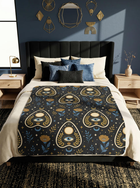 A rectangular velveteen minky blanket featuring a repeating dark academia pattern on a deep black background. The design includes gold and cream Ouija planchettes, each containing a blue all-seeing eye, a cream circle, and a light blue crescent moon framed by botanical leaves. Scattered between the planchettes are gold flowers with light blue stems, additional crescent moons, and small four-point stars. The reverse side of the blanket is made of a fluffy, textured off-white sherpa fleece.