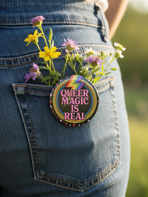 A round metal pinback button badge featuring a black background with the words QUEER MAGIC IS REAL in capital pink retro serif typography. A bright rainbow colored lightning bolt strikes from the top center, surrounded by concentric rainbow rings in red, orange, yellow, green, blue, and purple. The design is accented with small pink four-point stars and red crescent moons. The artwork features a realistic 2-D faux embroidery texture. The back has a silver metal safety pin clasp fastening.