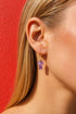 A close-up view of a person's ear wearing a pair of purple mushroom-shaped earrings against a red background.