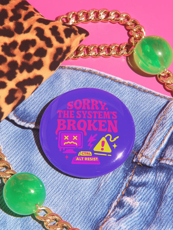 A vibrant purple pinback button with a glossy finish. The main text reads "SORRY, THE SYSTEM'S BROKEN" in a chunky, dripping pink font. Below the text are several retro tech icons in pink and yellow. On the left is a glitching 90s-style computer monitor with a sad face and X's for eyes. In the center is a yellow warning sign with an exclamation point, a lightning bolt, and sparkles. Below is a keyboard command parody that reads "CTRL ALT RESIST.