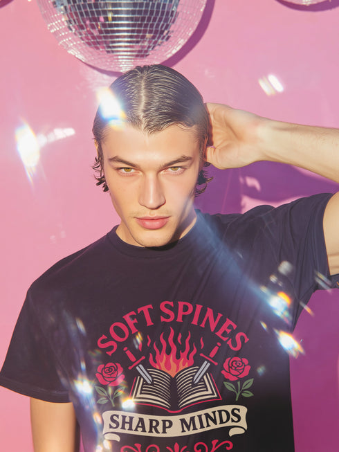 A black unisex t-shirt is shown flat on a neutral surface. The front of the shirt features a detailed graphic in a traditional tattoo style with red and white elements. In the center is an open book with flames rising from its pages. Two daggers pierce the book from either side. The book is flanked by two red roses with green leaves. Above the book, the words "SOFT SPINES" are written in a red arched font. Below, a white banner reads "SHARP MINDS." The overall aesthetic is gothic literary.