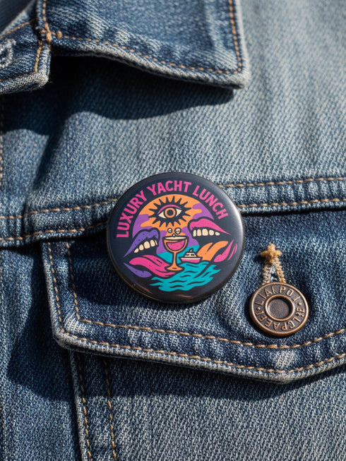 A round 1.75 inch glossy pinback button featuring satirical psychedelic artwork on a black background. The text LUXURY YACHT LUNCH arches over the top in bold neon pink letters. The central design depicts a surreal scene with a stylized third-eye sun, large open mouths with tongues sticking out, a smiling cocktail glass character, and a small white yacht floating on wavy teal water. The color palette includes retro 70s neon pink, orange, purple, and teal. The button has a safety pin backing.