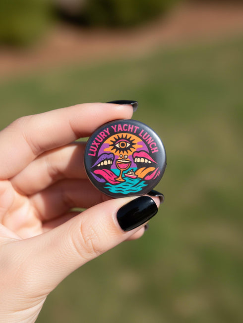 A round 1.75 inch glossy pinback button featuring satirical psychedelic artwork on a black background. The text LUXURY YACHT LUNCH arches over the top in bold neon pink letters. The central design depicts a surreal scene with a stylized third-eye sun, large open mouths with tongues sticking out, a smiling cocktail glass character, and a small white yacht floating on wavy teal water. The color palette includes retro 70s neon pink, orange, purple, and teal. The button has a safety pin backing.