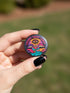 A round 1.75 inch glossy pinback button featuring satirical psychedelic artwork on a black background. The text LUXURY YACHT LUNCH arches over the top in bold neon pink letters. The central design depicts a surreal scene with a stylized third-eye sun, large open mouths with tongues sticking out, a smiling cocktail glass character, and a small white yacht floating on wavy teal water. The color palette includes retro 70s neon pink, orange, purple, and teal. The button has a safety pin backing.