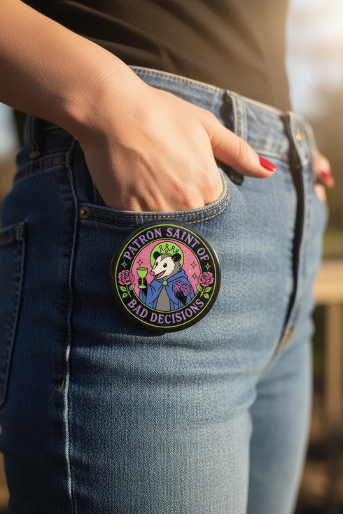 A round glossy metal pinback button featuring a flat 2D print illustration of an opossum wearing a blue cloak and a green crown. The opossum holds a neon green goblet in one paw and two purple dice in the other. A bright pink circle sits behind the opossum, surrounded by a black outer ring. Purple text reads PATRON SAINT OF BAD DECISIONS curved around the edge. Two pink roses with green leaves are on the sides. The design uses bold black outlines and bright neon colors.