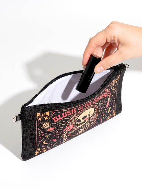 A rectangular gothic makeup bag made of black polyester with a visible woven linen-like crosshatch texture. The central illustration features a smiling human skull with flowing hair applying a pink heart-shaped blush to its cheek. Above the skull, the text BLUSH OF THE DAMNED appears. The design is framed by illustrations of lipstick tubes, mascara wands, bows. The bag includes a black zipper along the top edge and a removable black nylon wrist strap attached to the metal zipper pull.