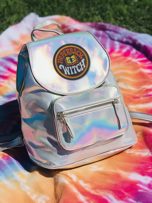 A round, black embroidered patch with a thick, textured merrow border stitched in alternating orange and yellow thread. In the center, the words "PAPERBACK WITCH" are embroidered in a retro 70s-style font. The word "PAPERBACK" is arched across the top in bold orange thread with a thin yellow outline. The word "WITCH" is across the bottom in puffy, bright white thread with a thin yellow outline. Between the words is a central icon of an open book, containing a stylized all-seeing eye.