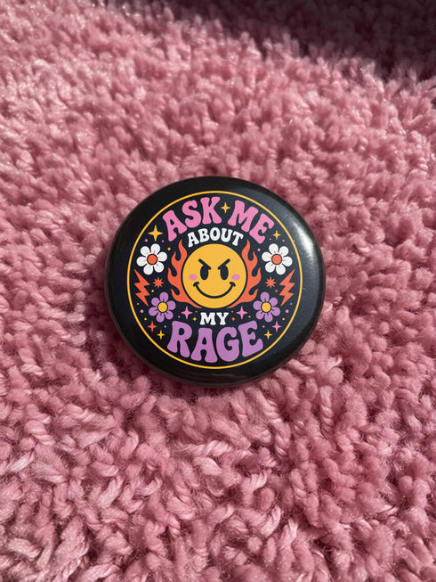 A round glossy metal pinback button featuring a flat 2D print design on a solid black background. In the center is an angry yellow smiley face surrounded by orange and red flames. Above the smiley face, pink text reads ASK ME and white text reads ABOUT. Below the face, white text reads MY and large purple text reads RAGE. The design is accented with two white daisies, two purple flowers, pink and orange four-point stars, and orange lightning bolts framing the outer edges.