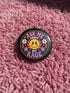A round glossy metal pinback button featuring a flat 2D print design on a solid black background. In the center is an angry yellow smiley face surrounded by orange and red flames. Above the smiley face, pink text reads ASK ME and white text reads ABOUT. Below the face, white text reads MY and large purple text reads RAGE. The design is accented with two white daisies, two purple flowers, pink and orange four-point stars, and orange lightning bolts framing the outer edges.