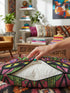 A square decorative pillow cover featuring a vibrant, symmetrical flat printed design that mimics the look of textured embroidery. Two black cats with large yellow eyes and pink noses face forward against a bright red background. Below the cats are large purple and pink flowers with green stems. Above the cats are green cactus shapes. The artwork uses bold, colorful outlines to create an optical illusion of stitching, though the fabric itself is smooth.