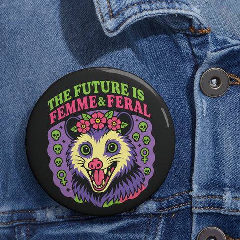 A round, black pinback button with a high-gloss finish features the text "THE FUTURE IS FEMME & FERAL" in a retro font of lime green and pastel pink. In the center, a screaming possum with wild eyes and a pink-lined mouth is illustrated in a pastel goth style with purple, yellow, and pink fur. The possum wears a pink flower crown. The illustration is framed by a circle of small skulls and pink Venus (female) symbols on a purple background. This is a piece of feminist possumcore flair.