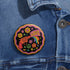 Round pinback button badge with a black background, featuring a retro illustration of a red revolver shooting out a rainbow flag that says “LOVE.” Surrounded by colorful 70s-style flowers and peace signs, outlined in wavy neon border. Glossy finish with vibrant pop colors and psychedelic design.