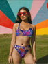 Psychedelic bikini bottom with multicolored tiger print on a neon leopard background; mid-rise cut with black lining and vibrant color blocking in purple, blue, pink, orange, and cream; made of recycled polyester and spandex blend with cheeky fit.