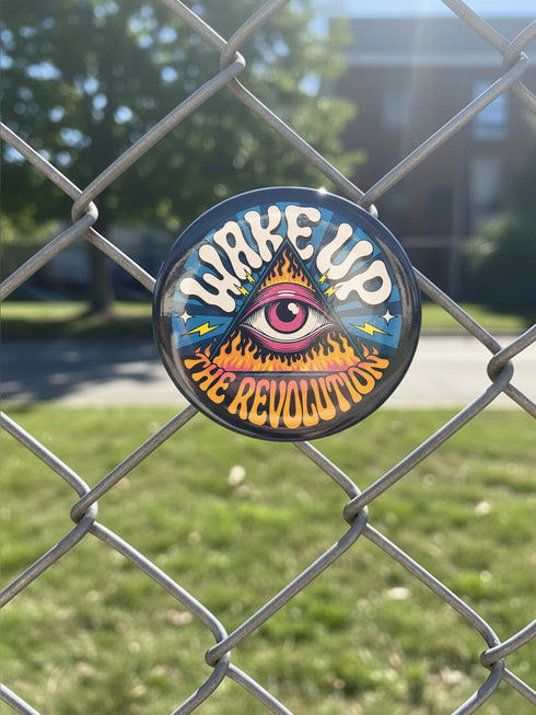 A round pinback button featuring a retro psychedelic design. The central focus is a stylized Eye of Providence in a pink triangle, surrounded by vibrant orange and yellow flames. Behind the triangle are teal and petrol blue sunburst rays. The text "WAKE UP" curves across the top in cream wavy letters, and "THE REVOLUTION" curves along the bottom in orange. The button has a glossy finish and is available in 1.75 inch or 2.25 inch sizes.