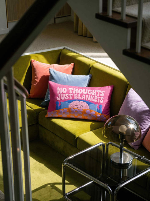 A bright magenta pink rectangular lumbar pillow cover featuring a flat 2D printed faux embroidery design. The printed illusion shows light pink bubble text reading NO THOUGHTS JUST BLANKETS. Below the text is a cartoon pink brain with a happy face, tucked into a blue bed with blue pillows. The brain is covered by a textured-looking orange blanket decorated with light purple daisies. The entire design is a smooth printed trompe l'oeil stitch effect, not real woven or stitched texture.