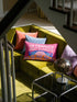 A bright magenta pink rectangular lumbar pillow cover featuring a flat 2D printed faux embroidery design. The printed illusion shows light pink bubble text reading NO THOUGHTS JUST BLANKETS. Below the text is a cartoon pink brain with a happy face, tucked into a blue bed with blue pillows. The brain is covered by a textured-looking orange blanket decorated with light purple daisies. The entire design is a smooth printed trompe l'oeil stitch effect, not real woven or stitched texture.