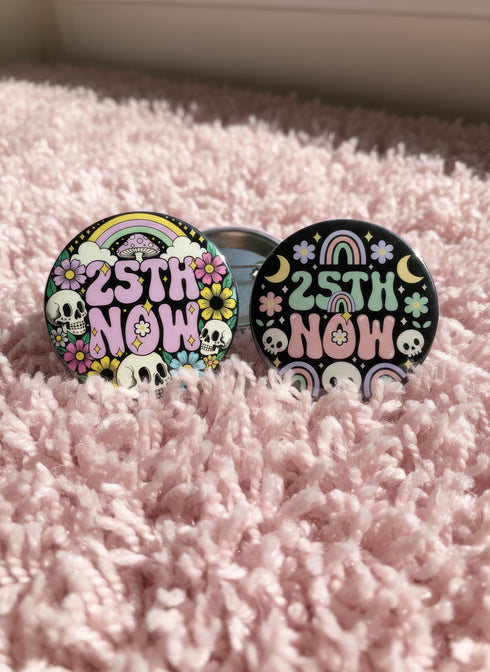 A round pinback button featuring a solid black background covered in a retro pastel colored design. The center reads 25TH NOW in a groovy bubbly font using pale pink and mint green referencing the 25th amendment. Surrounding the text is a maximalist cluster of cute but edgy motifs including smiling white skulls, pastel rainbows, pale yellow crescent moons, daisies, and a pink and white spotted mushroom. The button has a glossy laminate finish and a secure metal safety pin backing.