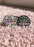 A round pinback button featuring a solid black background covered in a retro pastel colored design. The center reads 25TH NOW in a groovy bubbly font using pale pink and mint green referencing the 25th amendment. Surrounding the text is a maximalist cluster of cute but edgy motifs including smiling white skulls, pastel rainbows, pale yellow crescent moons, daisies, and a pink and white spotted mushroom. The button has a glossy laminate finish and a secure metal safety pin backing.