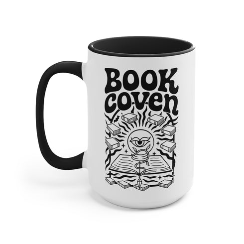 A two-tone ceramic mug with a glossy white exterior and a contrasting black interior, handle, and rim. The front of the mug features a black, rectangular graphic with the words "BOOK COVEN" in a bold, wavy, 70s-inspired retro font. Below the text is an occult-style illustration of an open book on a stand. An all-seeing eye with a snake coiled around it emerges from the book's pages, radiating lines of light. Smaller books float around the central image, completing the witchy, bookish design