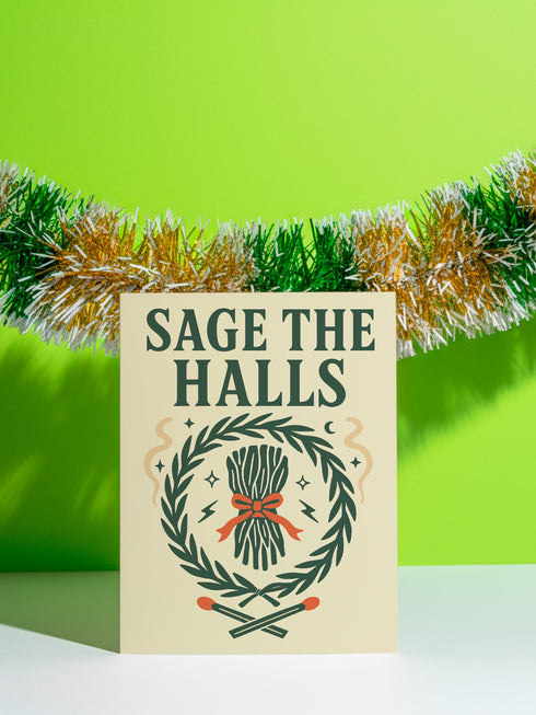 A detailed folk-art style illustration of a "Sage the Halls" holiday greeting card. The design features a bundle of sage smudge sticks tied with a red ribbon at the center of a dark green wreath. The wreath is accented with small crescent moons, stars, and smoke swirls. Below the wreath are two crossed matches with red tips. The card is printed on creamy beige paper with bold, dark green serif text. This funny spiritual and witchy Yule card is a unique pagan greeting for energy cleansing.
