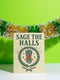 A detailed folk-art style illustration of a "Sage the Halls" holiday greeting card. The design features a bundle of sage smudge sticks tied with a red ribbon at the center of a dark green wreath. The wreath is accented with small crescent moons, stars, and smoke swirls. Below the wreath are two crossed matches with red tips. The card is printed on creamy beige paper with bold, dark green serif text. This funny spiritual and witchy Yule card is a unique pagan greeting for energy cleansing.