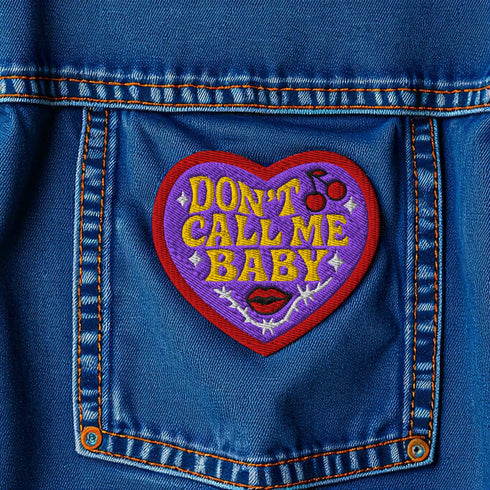 A heart-shaped embroidered patch with a thick red border and a vibrant purple background. In the center, the phrase "DON’T CALL ME BABY" is stitched in a bold, golden-yellow block font. The patch is decorated with small white sparkles, a pair of red cherries with a black musical note, a pair of red lips, and a grey thorny vine design below the text. The overall design has a rebellious, retro, punk aesthetic with a handmade, textured look.