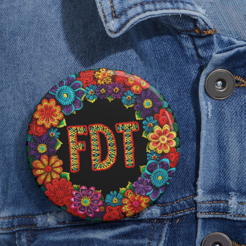 Circular black pinback button with bright 'FDT' lettering in orange/teal surrounded by colorful embroidery-style flowers in red, purple, blue, and yellow. Metal safety pin backing visible on reverse side. Political protest pin with vibrant Mexican folk art-inspired floral design.