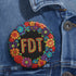 Circular black pinback button with bright 'FDT' lettering in orange/teal surrounded by colorful embroidery-style flowers in red, purple, blue, and yellow. Metal safety pin backing visible on reverse side. Political protest pin with vibrant Mexican folk art-inspired floral design.