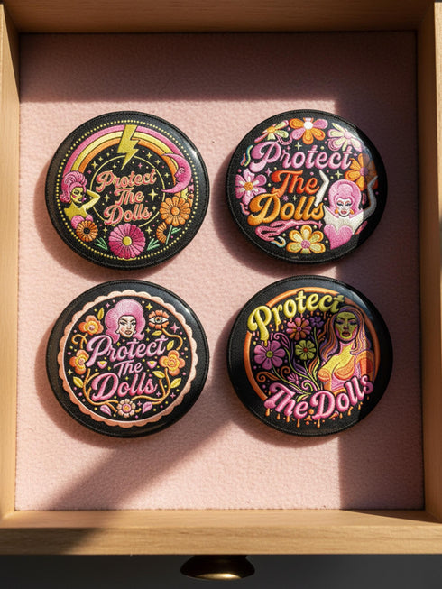 A circular black pinback button featuring the slogan Protect The Dolls in retro script. The design utilizes a high-fidelity trompe-l'œil print style that mimics the texture of raised thread embroidery with satin-stitch detailing. The artwork features vibrant hot pink, rust orange, and golden yellow motifs including drag queens with bouffant hair, 70s style daisies, lightning bolts, and a crescent moon with a face. The surface is smooth and glossy, creating a faux-textured illusion.