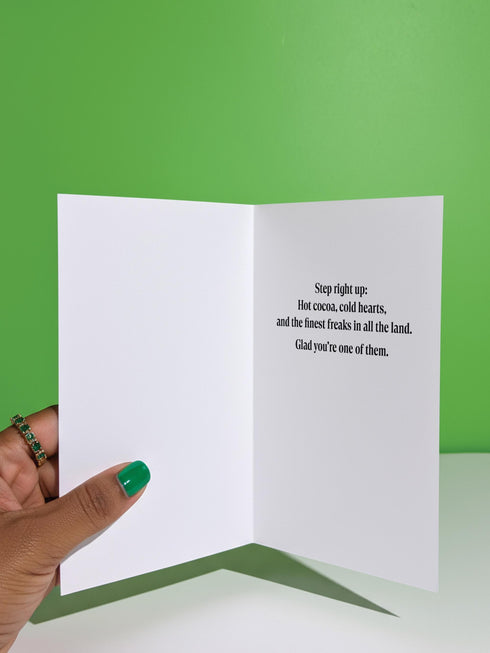 Maximalist Yuletide Freak Show 5x7 folded greeting card with a pearlescent shimmer finish. Features a vibrant, retro circus illustration in teal, pink, and yellow. The central motif is a bizarre, striped reindeer-tiger hybrid with a wind-up key, standing on a pedestal. The graphic border contains 'YULETIDE FREAK SHOW,' 'ODDITIES,' and 'WONDERS.' This unique and witty card is an unconventional alternative Christmas gift for oddity collectors.