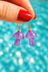 A hand holding a pair of purple mushroom-shaped earrings against a blurred blue background.