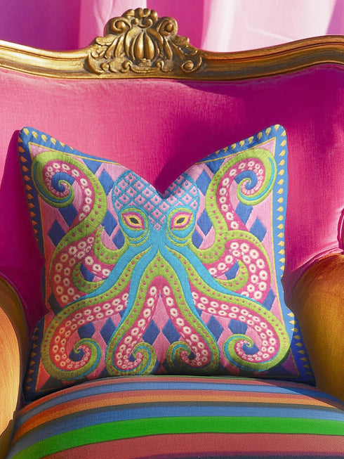 A square decorative throw pillow cover featuring a highly symmetrical bright octopus design. The octopus has a turquoise blue body with a diamond patterned head and eight curled tentacles detailed in bright pink and lime green. The background is a repeating diamond argyle pattern in royal blue and light pink. The artwork is a flat two dimensional print that mimics the textured look of intricate crewelwork embroidery. The pillow features a hidden zipper on the bottom edge. No insert included.