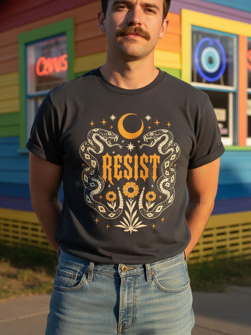 A unisex short sleeve graphic t shirt in dark charcoal black featuring a highly detailed symmetrical medallion print on the chest. In the center, the word RESIST is printed in a bold yellow gothic blackletter font. Two coiled white snakes with fine black scale details frame the text on the left and right. Above the text, a yellow crescent moon is flanked by white and yellow starbursts. Beneath, three yellow marigold flowers sit atop white stems and leafy foliage.