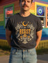 A unisex short sleeve graphic t shirt in dark charcoal black featuring a highly detailed symmetrical medallion print on the chest. In the center, the word RESIST is printed in a bold yellow gothic blackletter font. Two coiled white snakes with fine black scale details frame the text on the left and right. Above the text, a yellow crescent moon is flanked by white and yellow starbursts. Beneath, three yellow marigold flowers sit atop white stems and leafy foliage.