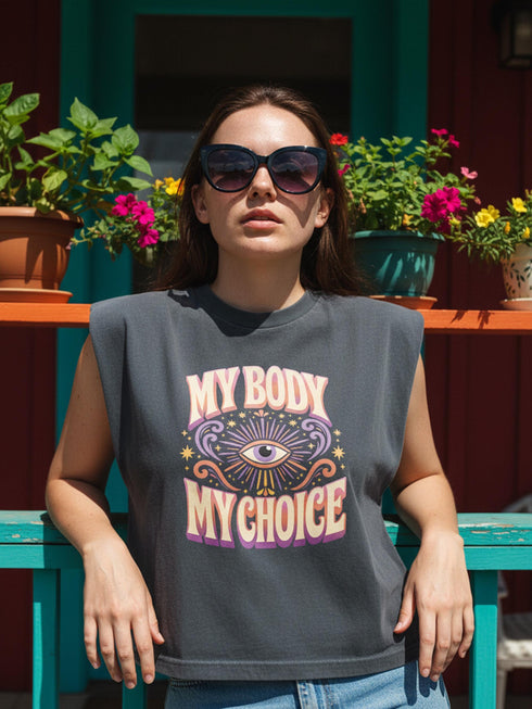 A garment dyed charcoal grey women's muscle tee featuring a slightly cropped boxy fit with structured shoulders and a wide rib collar. The front displays a retro 70s psychedelic graphic printed in purple, orange, and yellow. The text reads MY BODY MY CHOICE surrounding a central eye motif with a radial sunburst, framed by symmetrical mystical swirls and four point stars. The vintage style cotton shirt offers a faded aesthetic, presented on its own without background lifestyle elements.