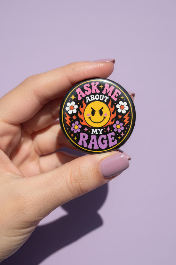 A round glossy metal pinback button featuring a flat 2D print design on a solid black background. In the center is an angry yellow smiley face surrounded by orange and red flames. Above the smiley face, pink text reads ASK ME and white text reads ABOUT. Below the face, white text reads MY and large purple text reads RAGE. The design is accented with two white daisies, two purple flowers, pink and orange four-point stars, and orange lightning bolts framing the outer edges.