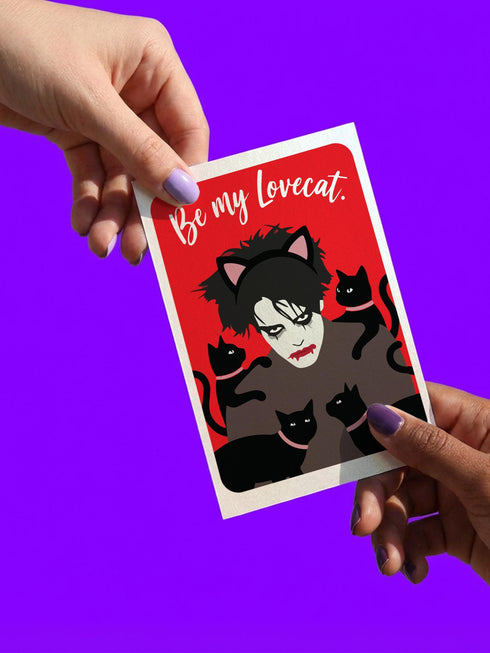 A vertical greeting card featuring a flat vector illustration of Robert Smith from The Cure against a vibrant red background. He has messy black hair, pale skin with smeared lipstick, and wears black cat ears. He is surrounded by four black cats with pink collars. White script text at the top reads "Be my Lovecat." The card is printed on shimmery pearlescent cardstock that catches the light. A high-quality alternative Valentine or anniversary card for goth music lovers and post-punk fans.