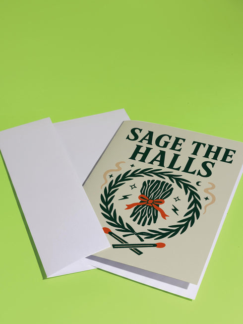 A detailed folk-art style illustration of a "Sage the Halls" holiday greeting card. The design features a bundle of sage smudge sticks tied with a red ribbon at the center of a dark green wreath. The wreath is accented with small crescent moons, stars, and smoke swirls. Below the wreath are two crossed matches with red tips. The card is printed on creamy beige paper with bold, dark green serif text. This funny spiritual and witchy Yule card is a unique pagan greeting for energy cleansing.