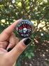 A collection of four glossy black circular pinback buttons featuring striking red and white gothic illustrations. One button shows "Midnight Graveyard Society" text with bats and tombstones. Another reads "Cemetery Picnic Society" with a coffin and umbrella icon. A third features a detailed white skull and red roses with "Memento Mori" text below. The final pin displays "Nocturnal Flower Society" surrounded by floral vines.