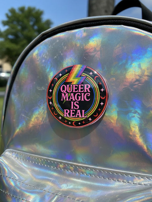 A round metal pinback button badge featuring a black background with the words QUEER MAGIC IS REAL in capital pink retro serif typography. A bright rainbow colored lightning bolt strikes from the top center, surrounded by concentric rainbow rings in red, orange, yellow, green, blue, and purple. The design is accented with small pink four-point stars and red crescent moons. The artwork features a realistic 2-D faux embroidery texture. The back has a silver metal safety pin clasp fastening.