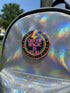 A round metal pinback button badge featuring a black background with the words QUEER MAGIC IS REAL in capital pink retro serif typography. A bright rainbow colored lightning bolt strikes from the top center, surrounded by concentric rainbow rings in red, orange, yellow, green, blue, and purple. The design is accented with small pink four-point stars and red crescent moons. The artwork features a realistic 2-D faux embroidery texture. The back has a silver metal safety pin clasp fastening.