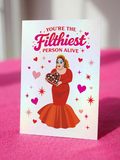 A 5x7 inch pearlescent silver greeting card featuring a flat vector illustration of the drag queen Divine in a red mermaid gown holding a heart-shaped box of chocolates. The text above reads "You're The Filthiest Person Alive" in bold red script with pink sparkles. The card has a shimmering finish that reflects light. Inside text reads "...and I love it." The back features the Honey Dazed logo. Includes a matching envelope.