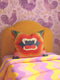 a vibrant and colorful bedroom scene, with a yellow headboard and a pillow adorned with a bold, graphic design. The pillow has a mouth and teeth motif, adding a playful and whimsical touch to the room's decor. The bed is covered in a colorful striped blanket, complementing the overall lively atmosphere of the space.