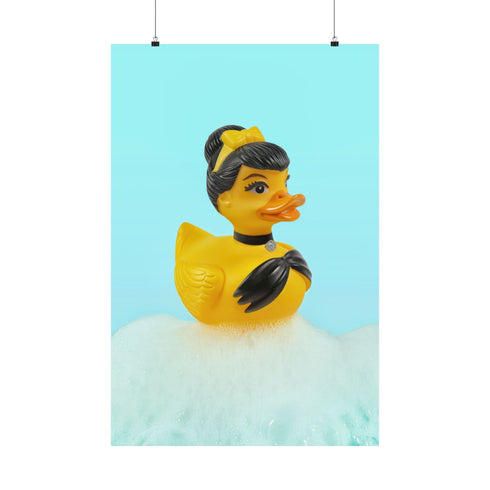A chic rubber duck styled like Audrey Hepburn’s Holly Golightly, wearing a high bun, yellow bow, black gloves, and a sparkly choker. The duck floats in a pile of foam and is framed in white on a bubbly bathroom shelf with a red tile backdrop and soap dispenser.