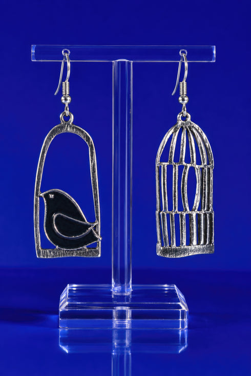 A pair of silver earrings on a clear stand, with one earring designed to resemble a bird in a cage and the other earring depicting a bird in an open cage.