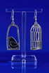 A pair of silver earrings on a clear stand, with one earring designed to resemble a bird in a cage and the other earring depicting a bird in an open cage.