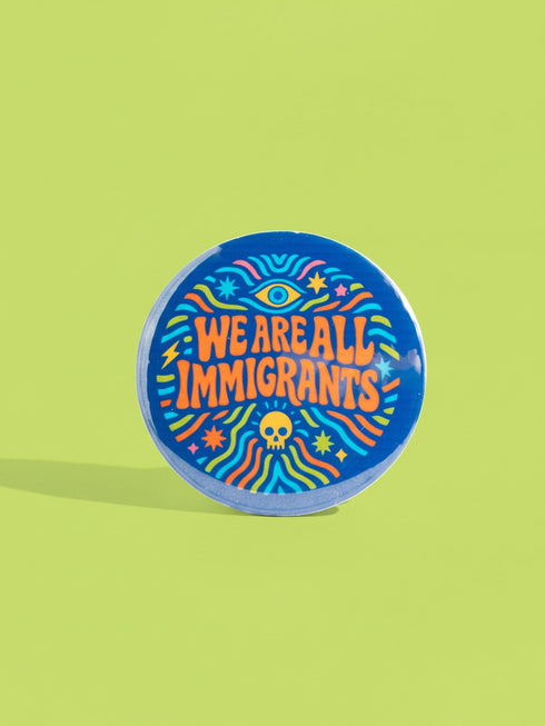 A round 1.75 inch blue pinback button featuring retro psychedelic artwork. The text reads We Are All Immigrants in a groovy orange and red 1970s font style centered on the badge. A mystic third eye sits above the text while a small skull sits below it. Wavy rainbow lines in cyan, green, and orange radiate outward from the center along with small starbursts and lightning bolts. The button has a glossy finish reflecting light.