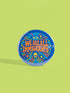 A round 1.75 inch blue pinback button featuring retro psychedelic artwork. The text reads We Are All Immigrants in a groovy orange and red 1970s font style centered on the badge. A mystic third eye sits above the text while a small skull sits below it. Wavy rainbow lines in cyan, green, and orange radiate outward from the center along with small starbursts and lightning bolts. The button has a glossy finish reflecting light.