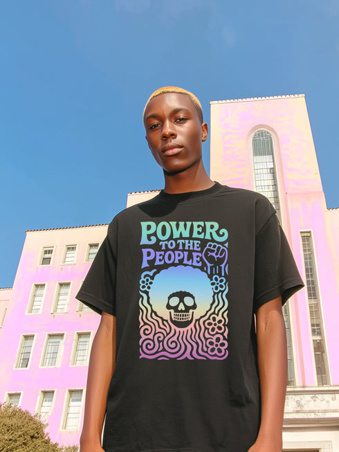 A detailed view of a black unisex graphic t-shirt made of soft cotton, shown flat. The shirt features a large, rectangular design on the chest inspired by 70s psychedelic protest art. The words "POWER TO THE PEOPLE" are printed at the top in a thick, rounded, groovy font. Below the text is a stylized human skull with a large, perfectly round afro hairstyle. The entire design, including the text, skull, and afro, is filled with a vibrant color gradient that shifts from deep purple to hot pink.